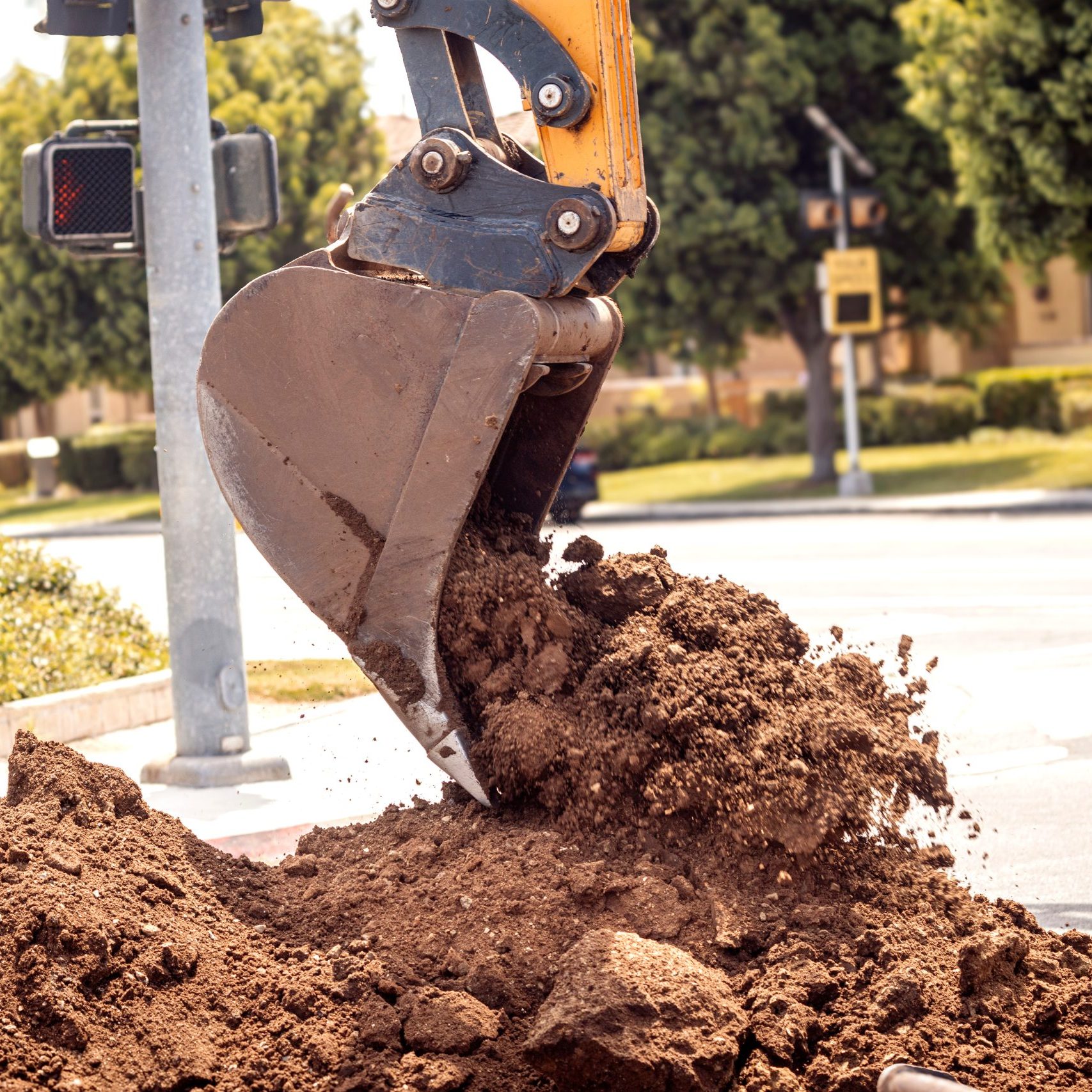 Close up of a backhoe scoop bucket filled with dirt  that is pouring out ontop a dirt pile.